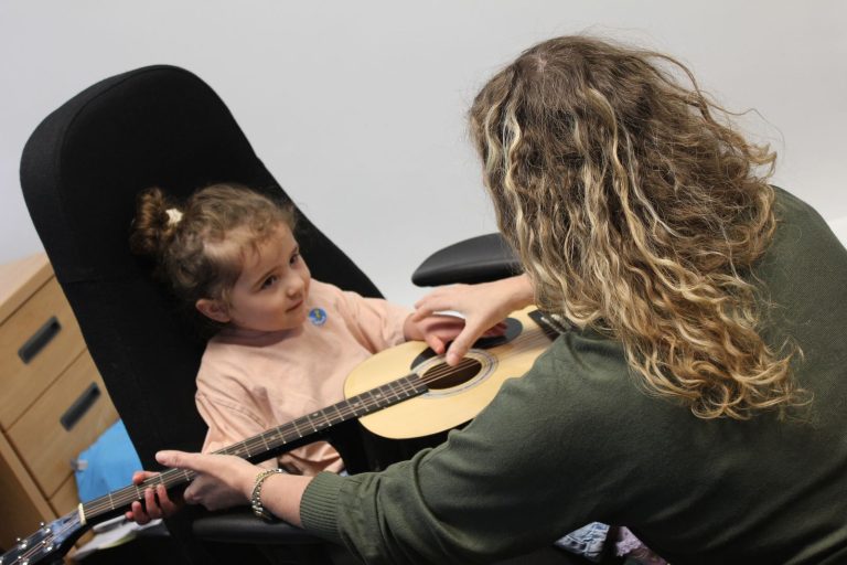 A child strumming a guitar with help from the music teacher