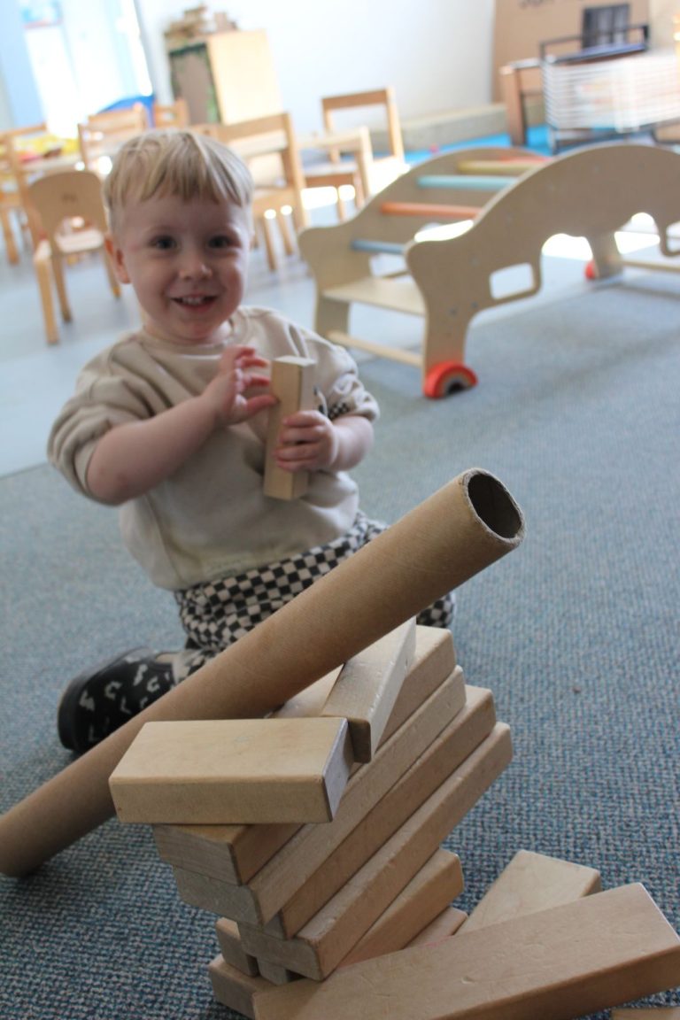 A smiling toddler building with cardboard tubes and wooden blocks