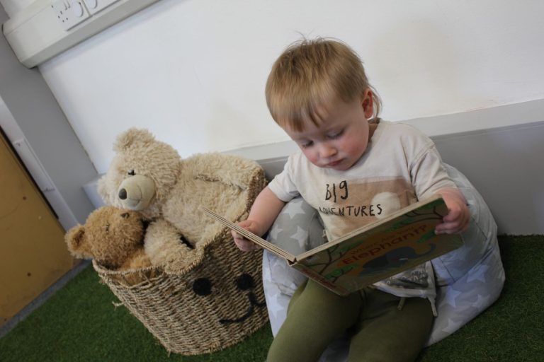 A baby reading a board book next to a basket of teddies
